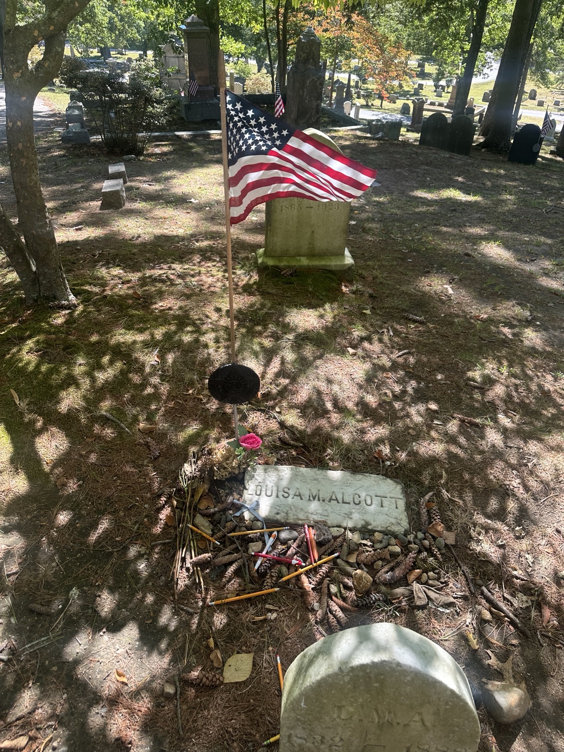 Louisa May Alcott’s grave on a wooded slope, with pencils, pinecones, a pink rose and an American flag in dappled sunlight.