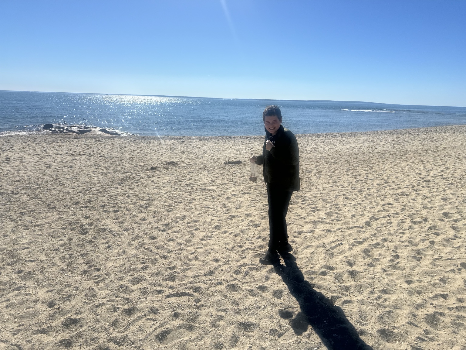 James smiling toward the camera on a broad sandy beach, with blue water and sky behind him.