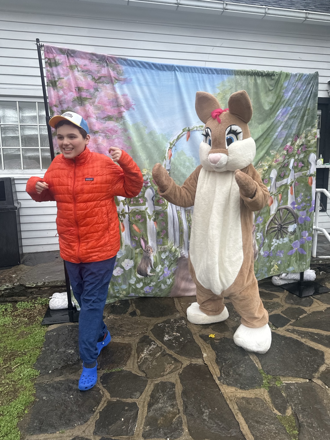 A teenager in an orange jacket smiles beside the Easter Bunny, with both raising their arms in front of a spring backdrop.