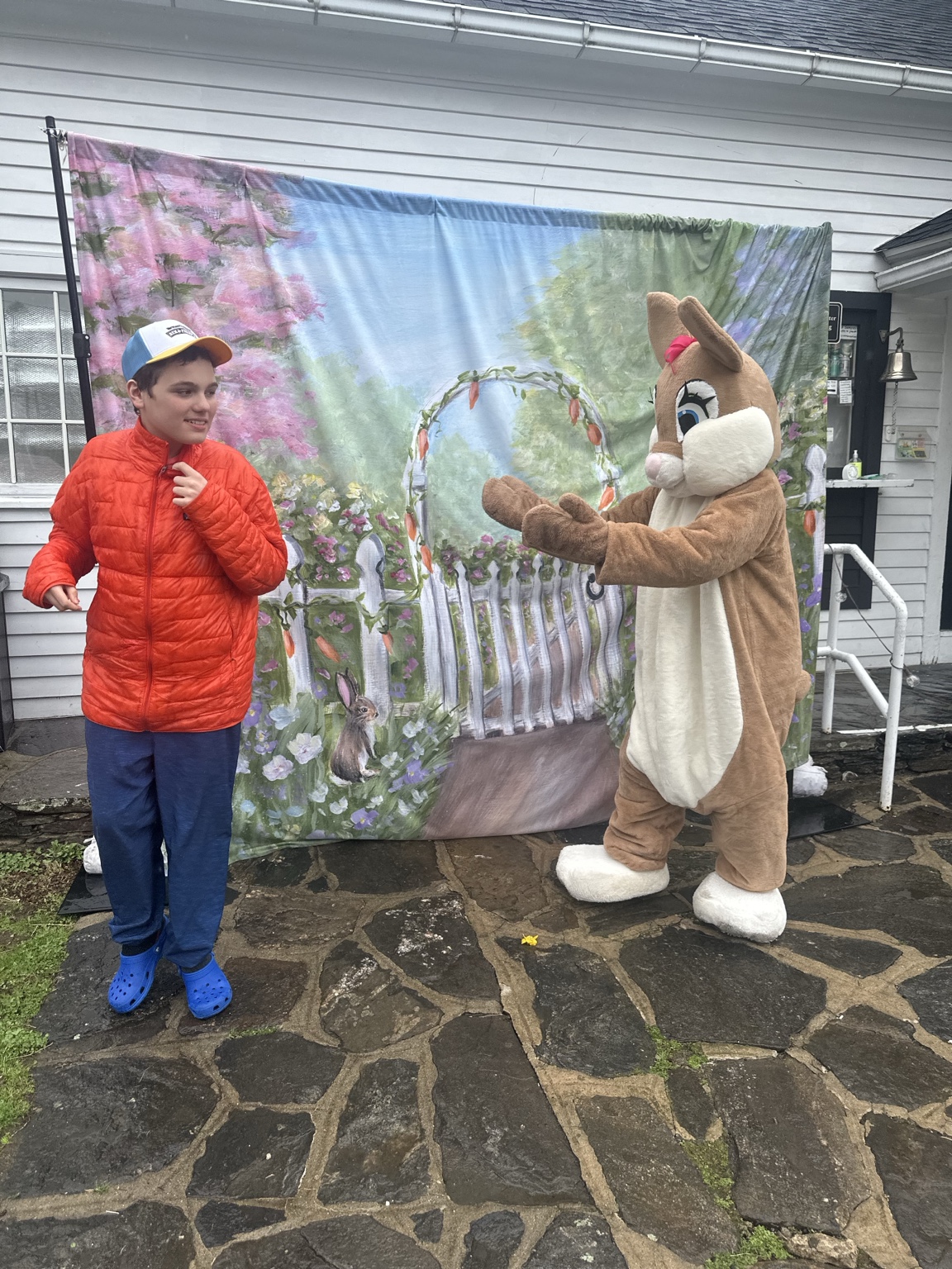 A teenager looks toward the Easter Bunny, who holds both paws out in front of a spring garden backdrop.