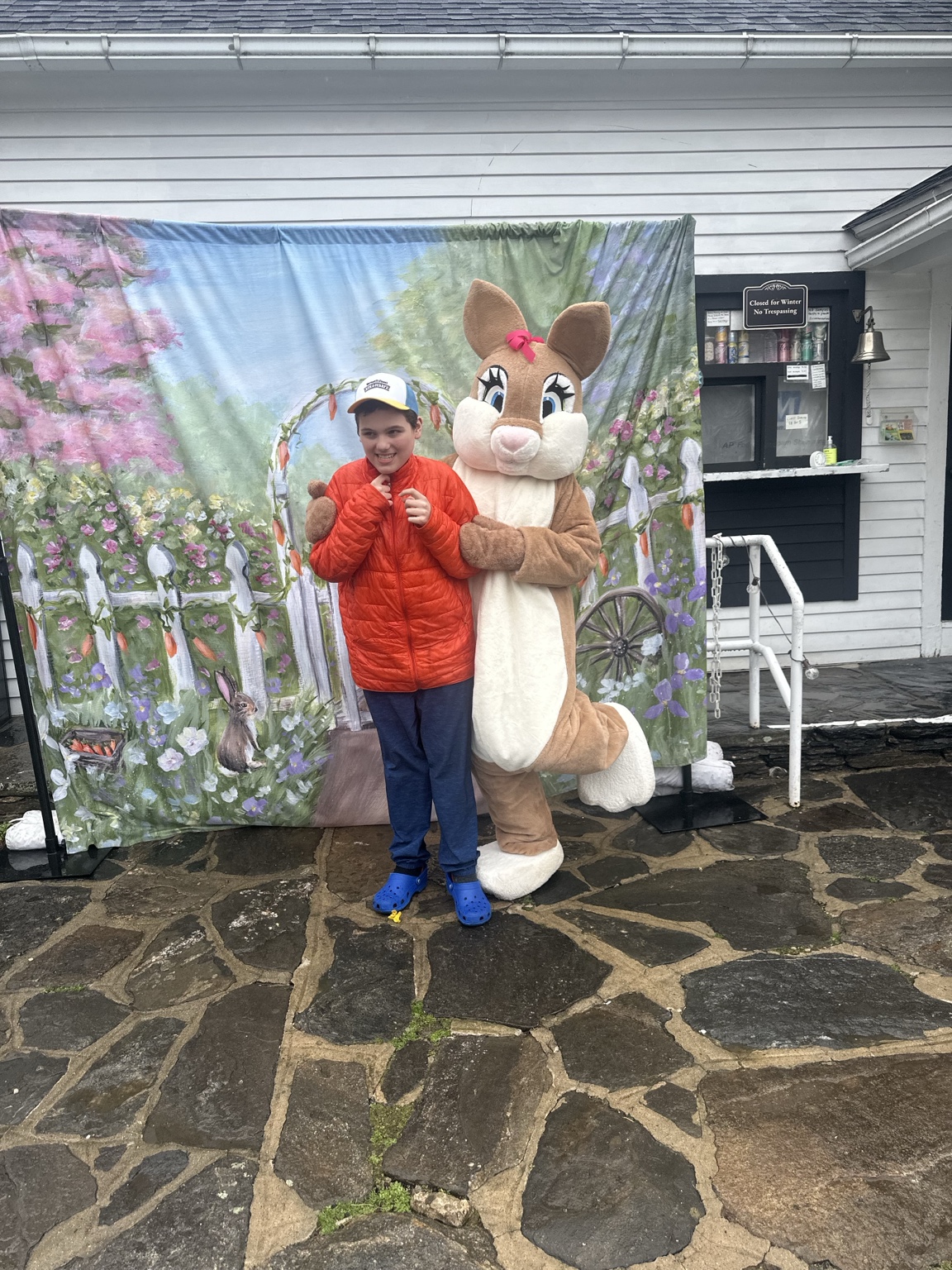 A teenager and the Easter Bunny pose side by side in front of a floral backdrop, with the bunny lifting one foot.