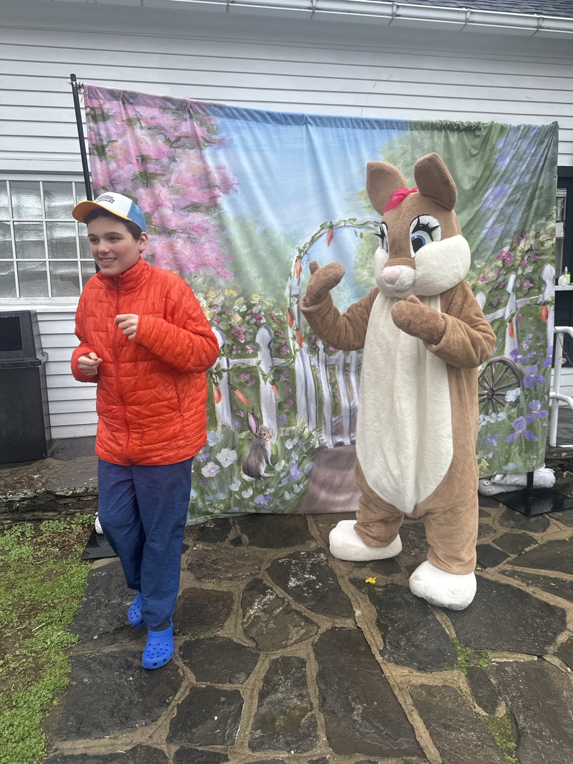 A teenager in an orange jacket stands beside a brown-and-cream Easter Bunny raising a paw.
