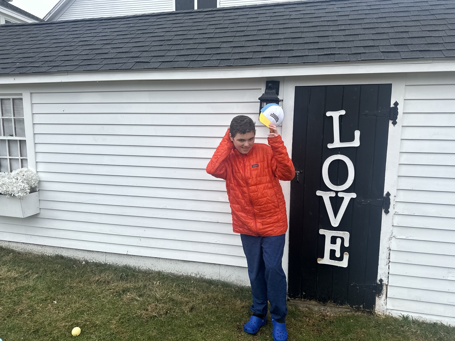 A teenager lifts a cap beside the black LOVE door of a white farm building, with a yellow egg on the grass.