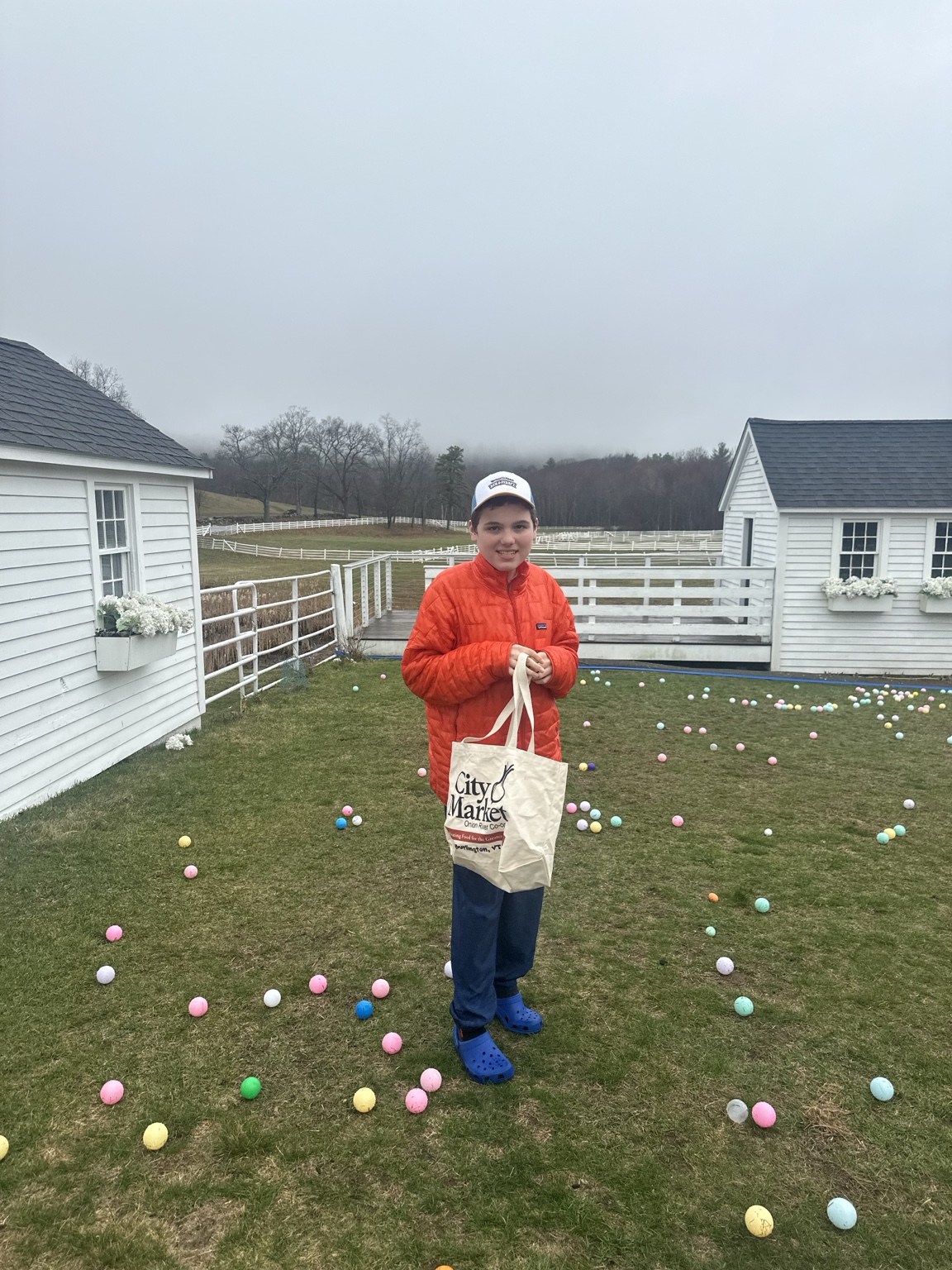 A teenager holds a canvas tote bag on a lawn scattered with colorful Easter eggs, between white farm buildings.