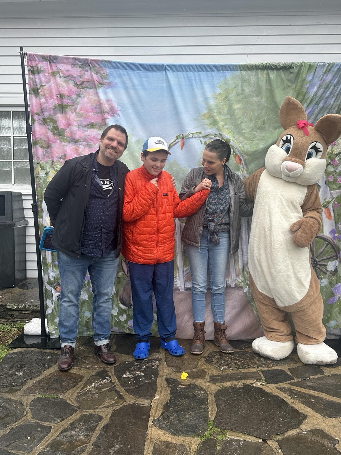 Three family members stand together beside the Easter Bunny on a stone patio in front of a floral backdrop.