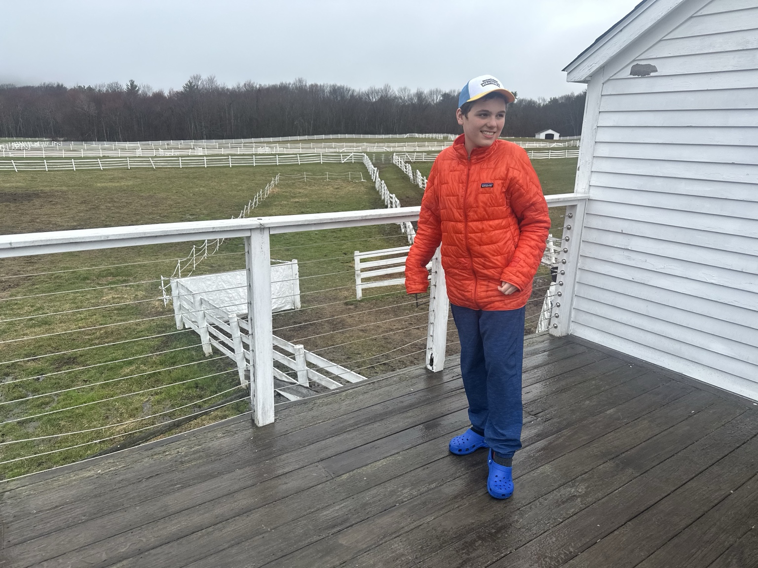 A teenager stands on a wooden deck overlooking green fields divided by rows of white fences.