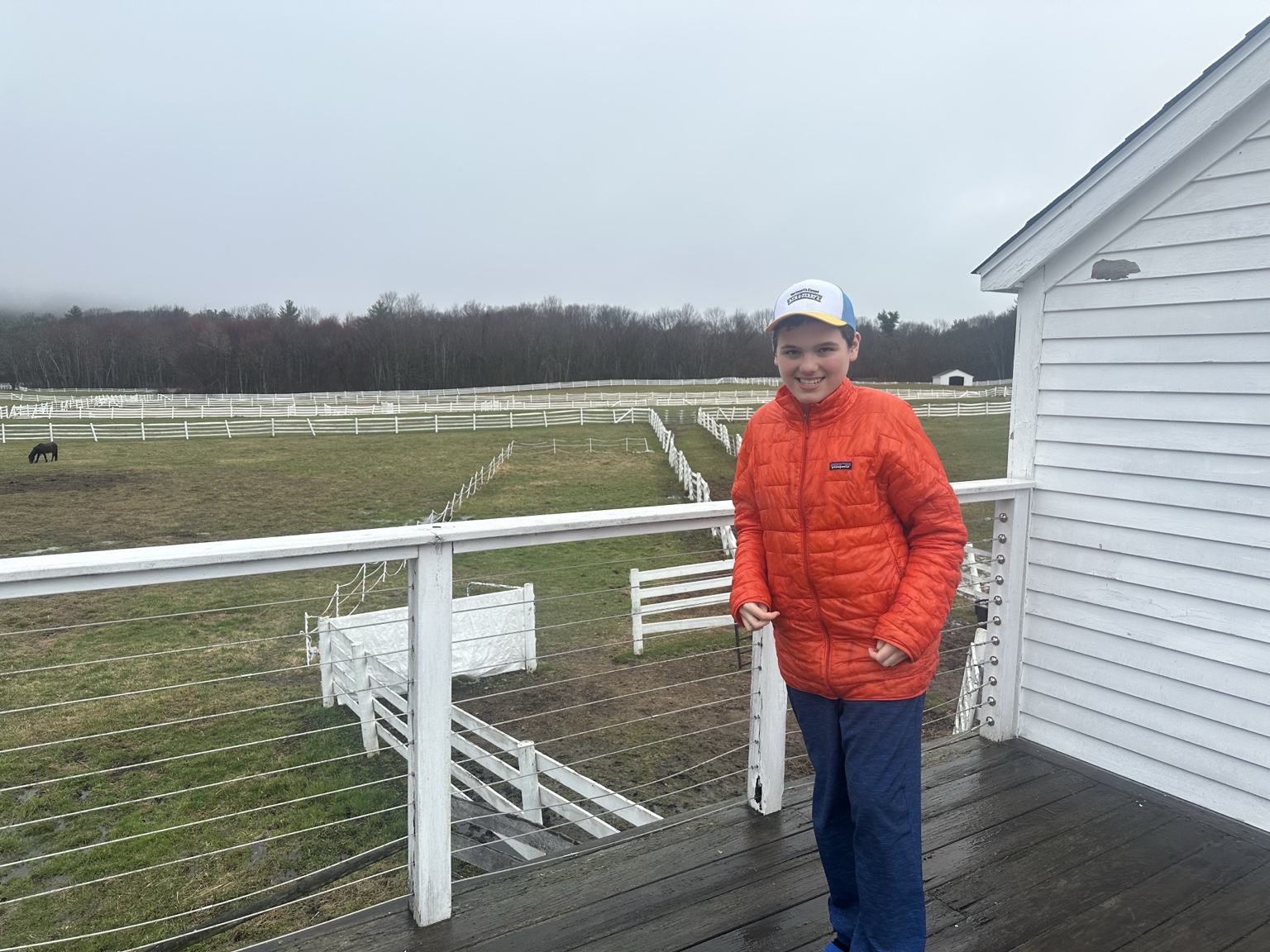 A teenager in an orange jacket stands on a wooden deck with white fences, open fields and a grazing horse in the distance.