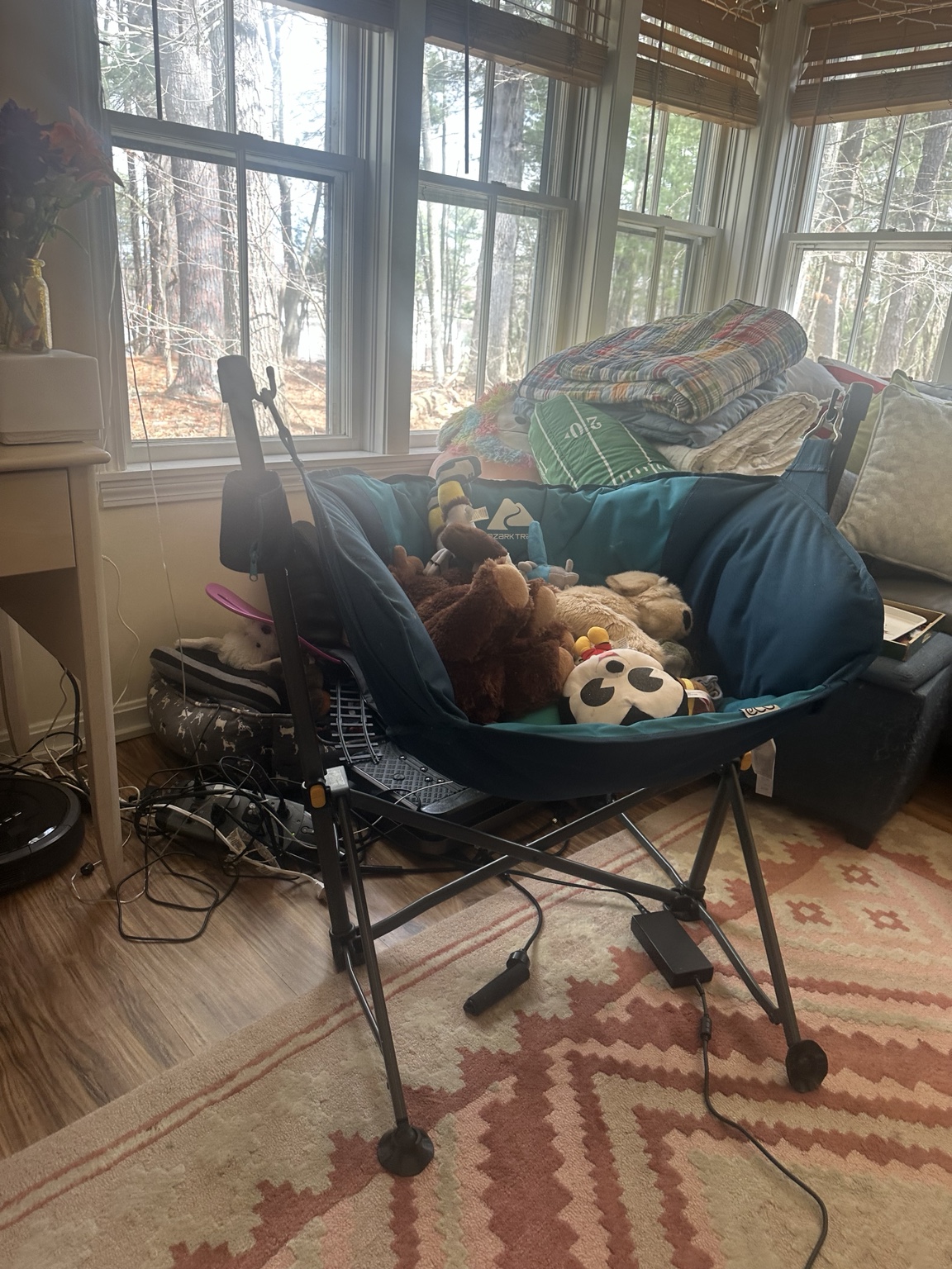 Blue hammock-style camping chair filled with stuffed animals beside tall sunroom windows, with folded quilts behind it.