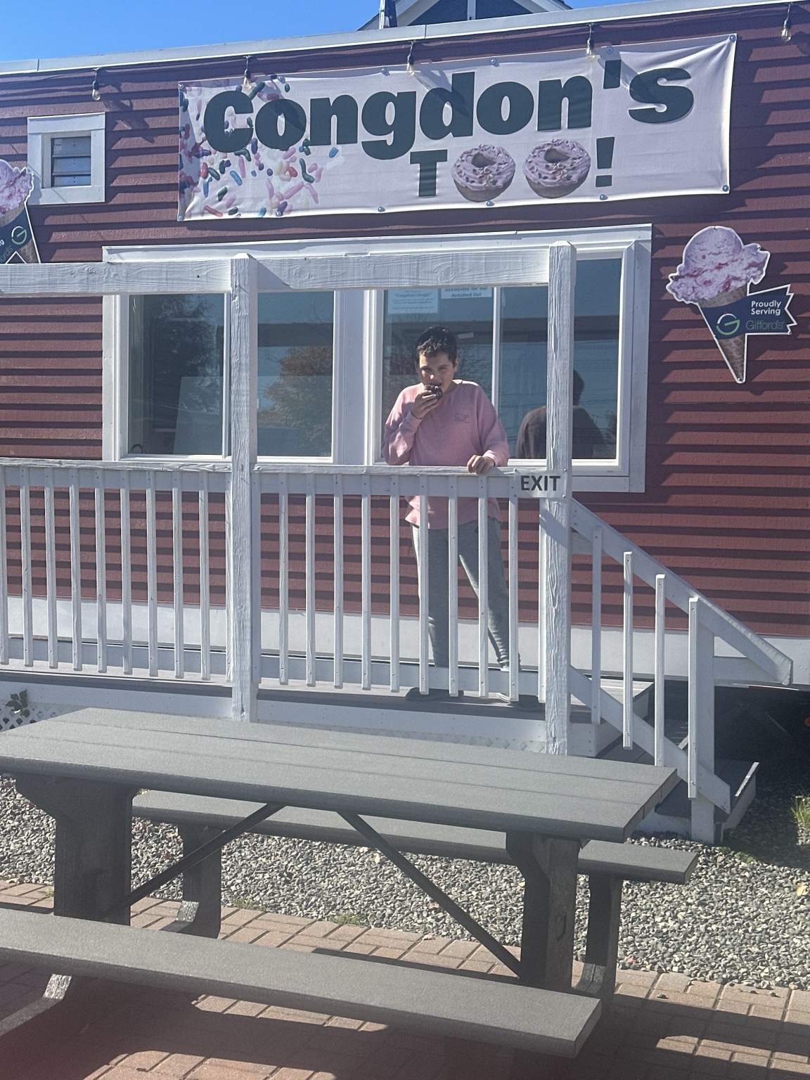James taking a bite of a doughnut on the raised Congdon’s Too porch, with an exit sign and picnic table nearby.
