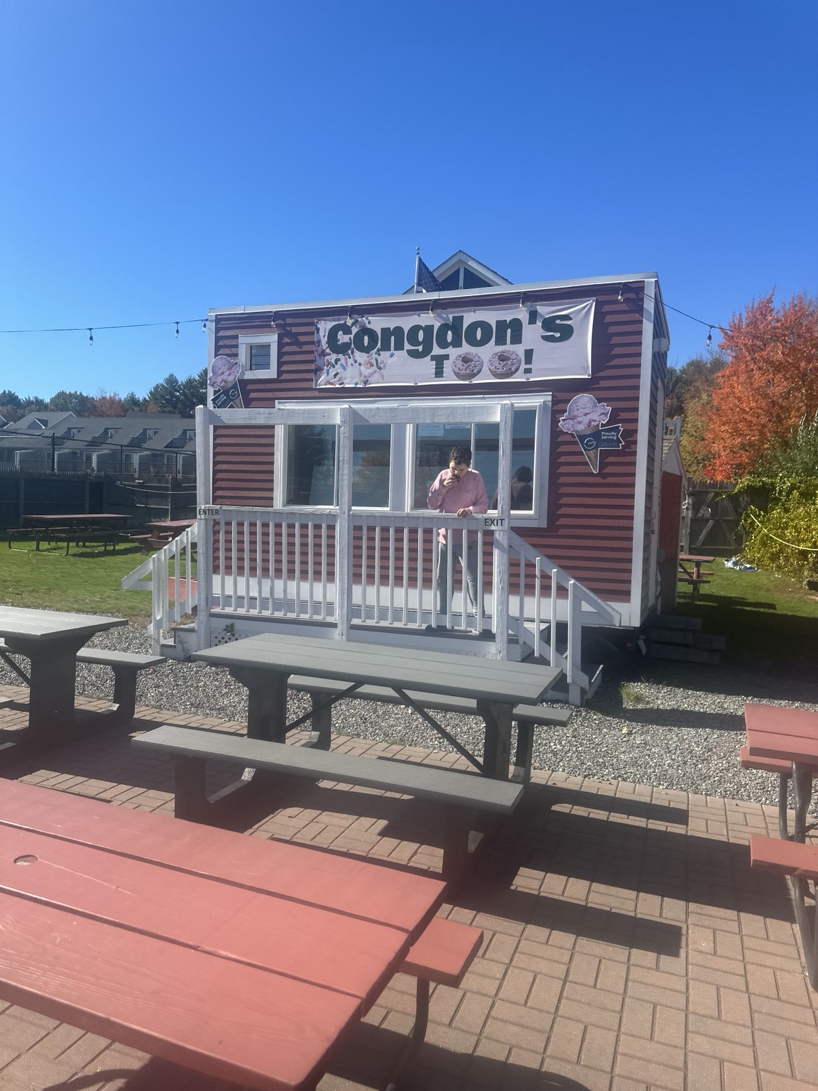 Congdon’s Too stand with entrance and exit steps, fixed-bench picnic tables on brick paving, and adjacent gravel and grass.