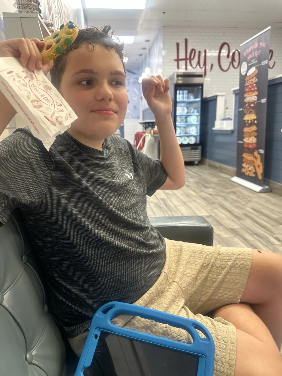 James sits comfortably indoors at Cookie Monstah, playfully holding a colorful cookie and its wrapper; his blue-cased iPad is nearby.