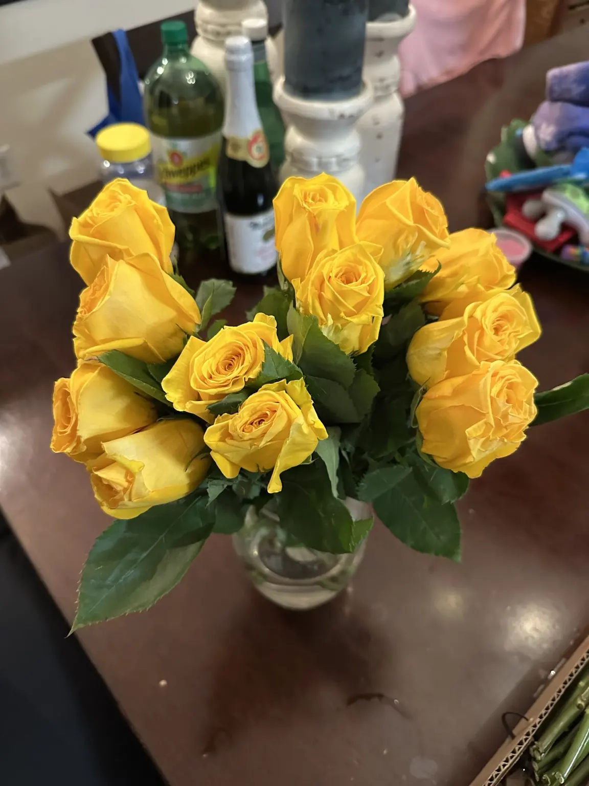 Yellow roses on a dark table with fidgets, bottles and everyday household things nearby.