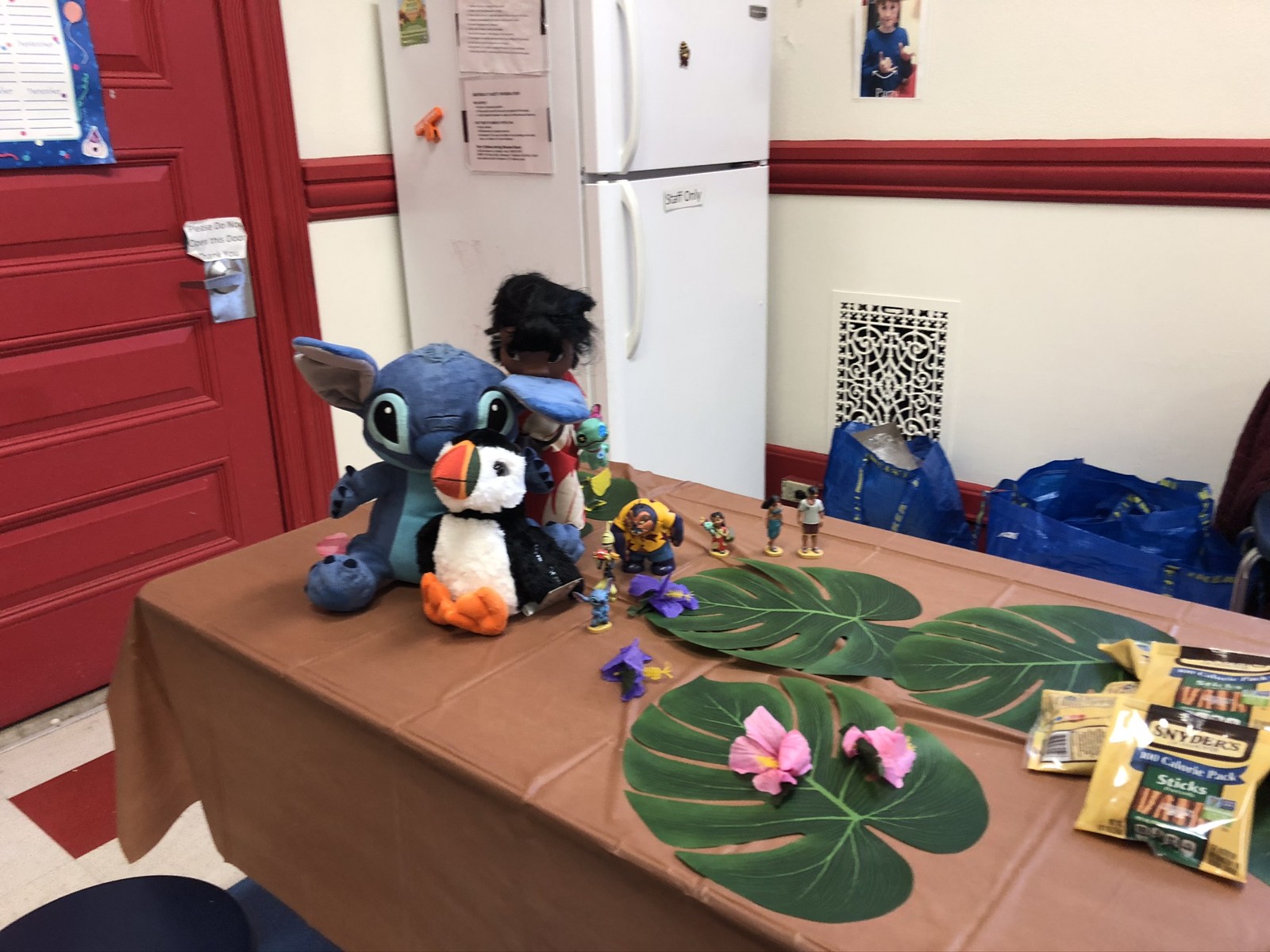 A blue Stitch plush beside a smaller white plush, paper leaves and flowers on a brown-covered table.