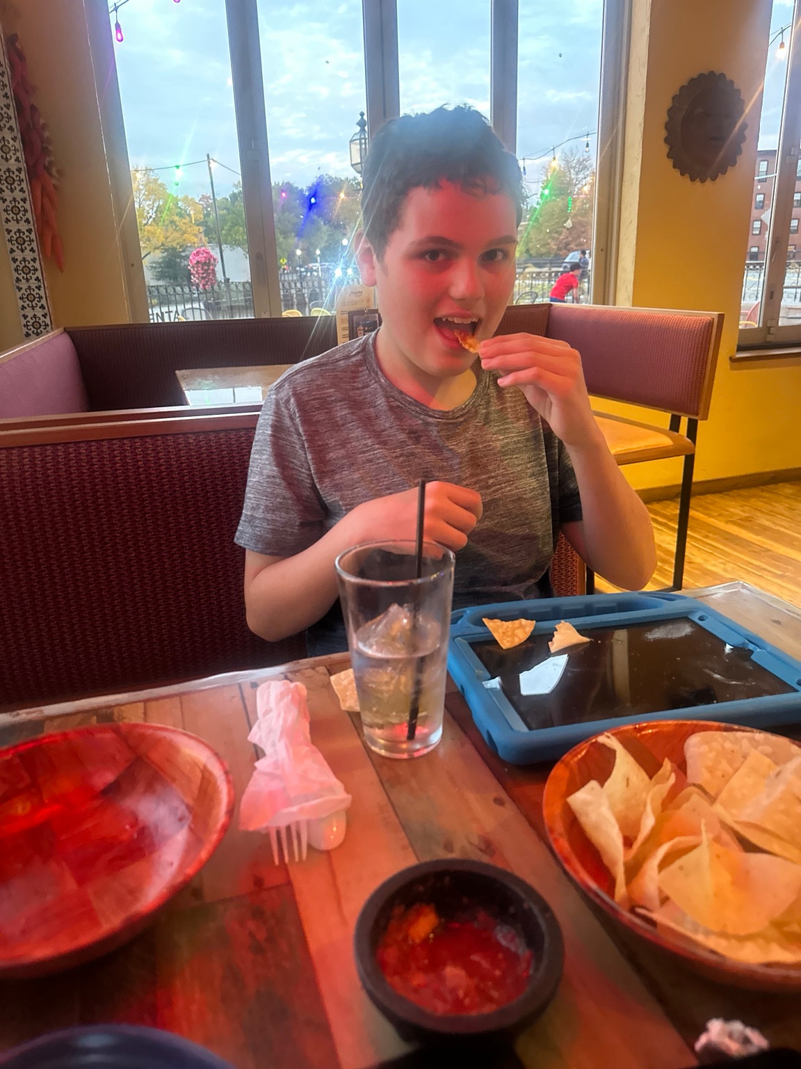 A diner eating a tortilla chip at a window-side table, with chips, salsa and a blue-cased tablet in front.