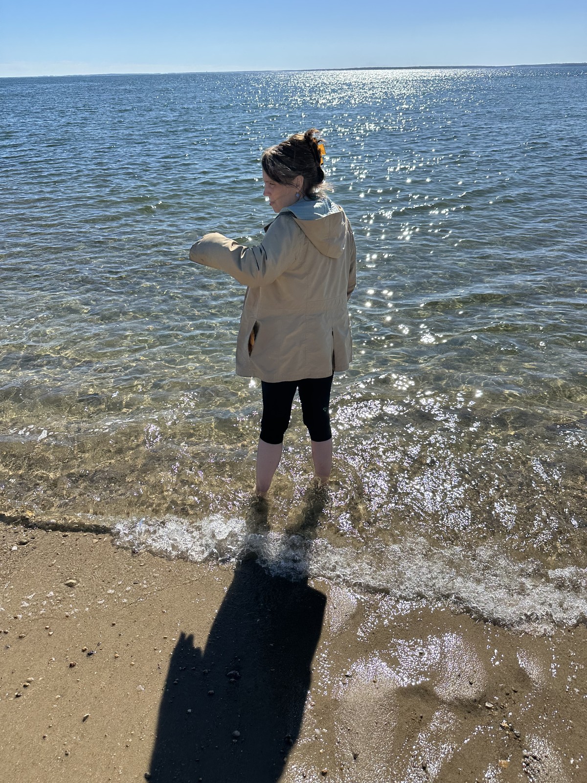 James’s mother turns toward the beach while standing barefoot in the sunlit shallows.