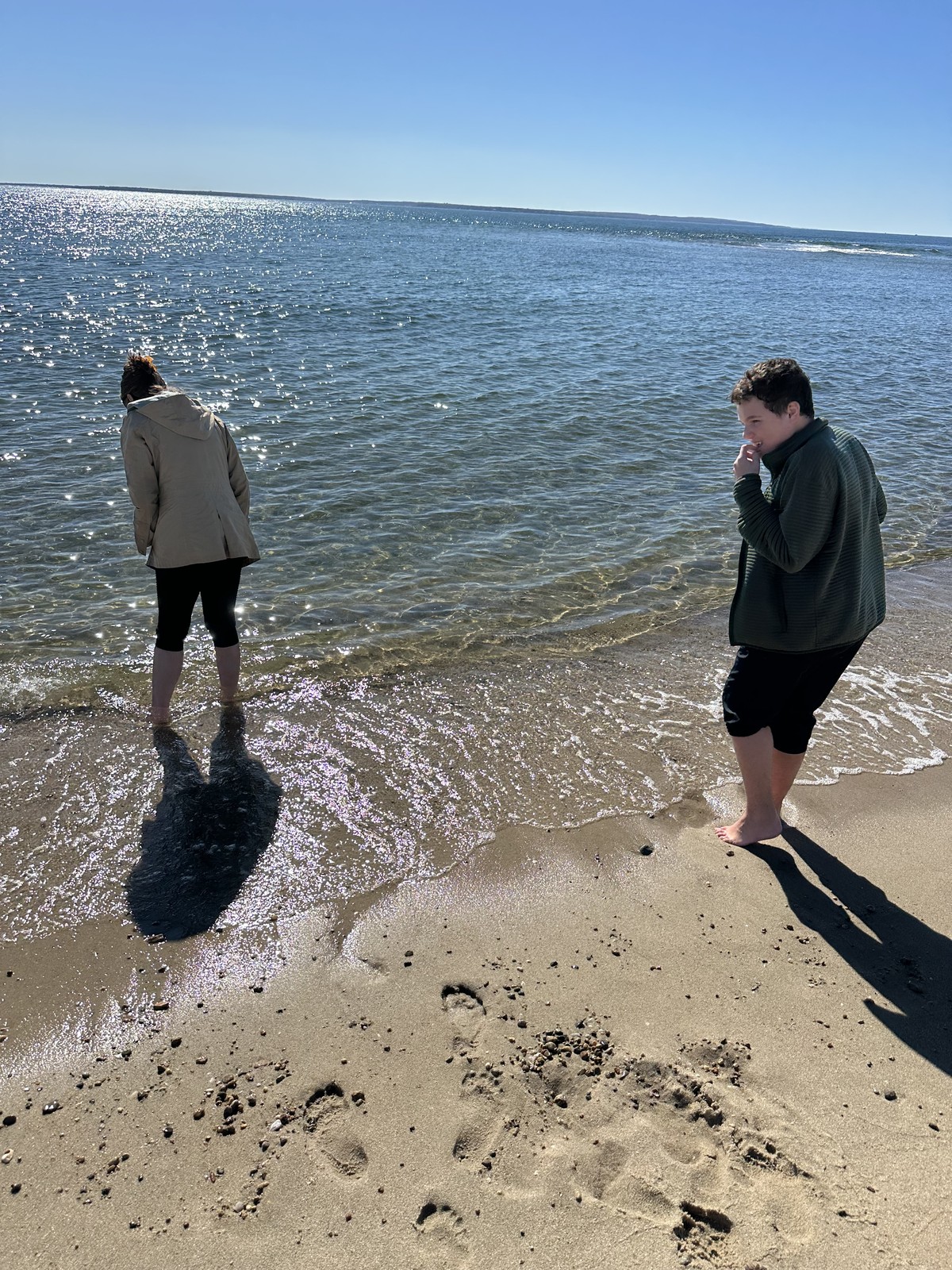 James and his mother stand beside one another at the water’s edge, with their shadows across the beach.