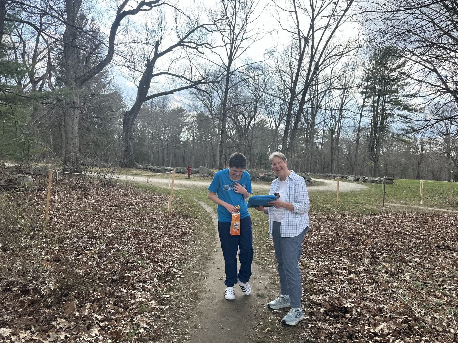 James and Susan laughing on a leaf-lined dirt path; James holds a bag of Goldfish and Susan holds a blue-cased tablet.