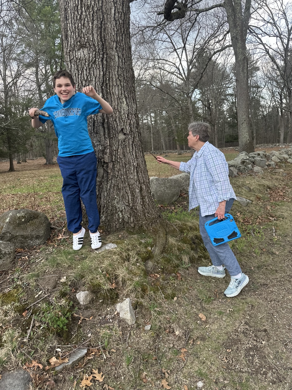 James smiling beside a large tree as Susan holds a twig toward its bark, with a blue-cased tablet in her other hand.