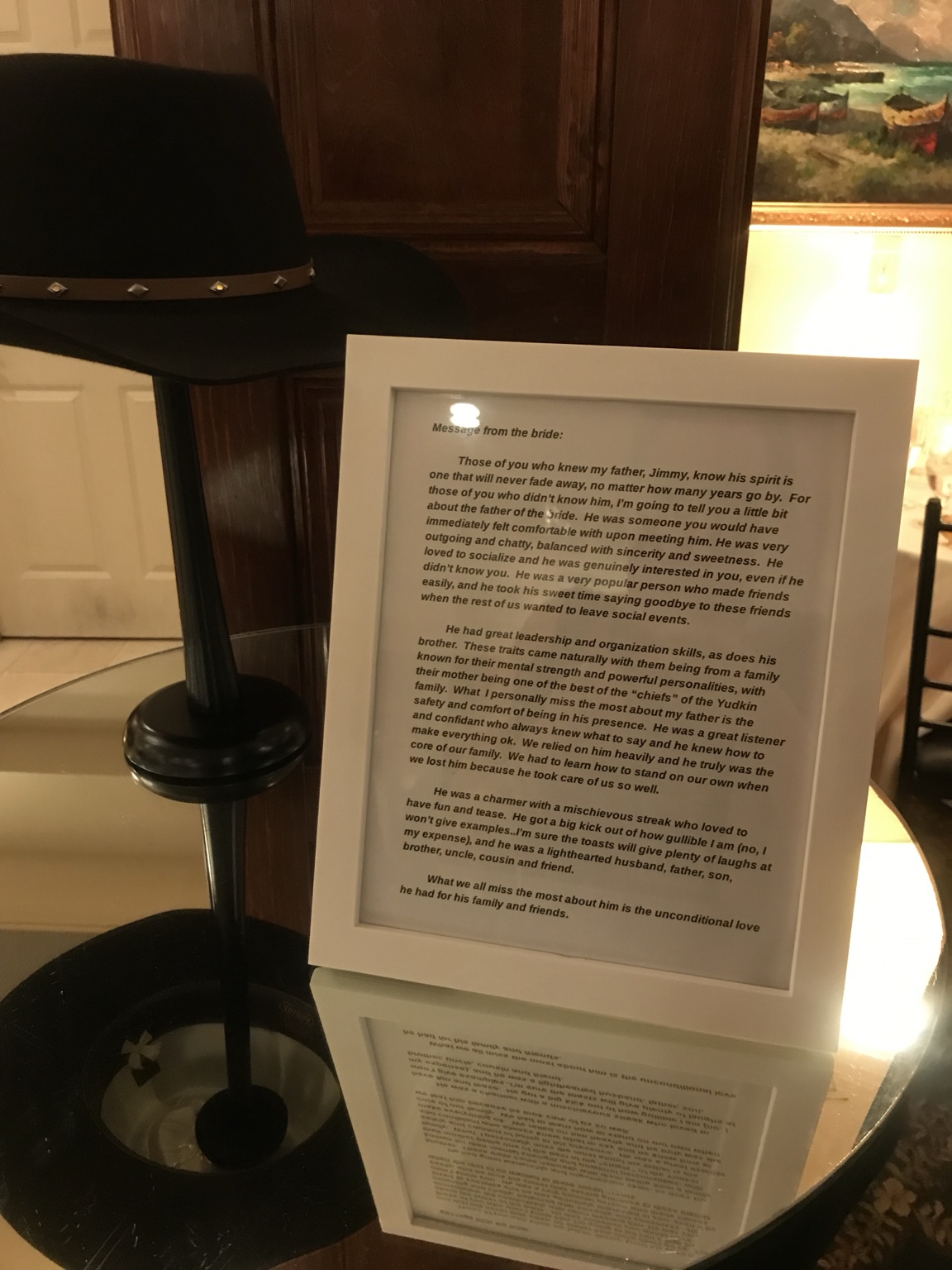 A framed message from the bride displayed on a round glass table beside a black hat.