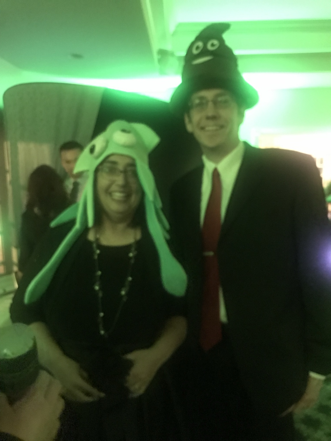 Two smiling wedding guests wearing playful oversized novelty hats in a green-lit reception room.