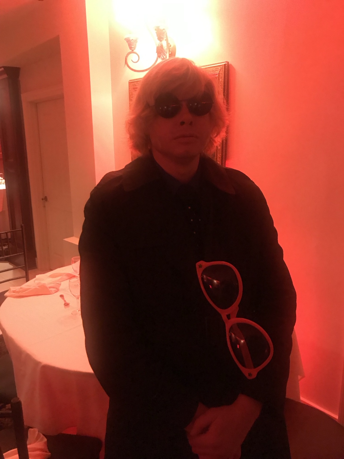 A guest in dark clothing and sunglasses holding oversized red sunglasses under red party lighting.