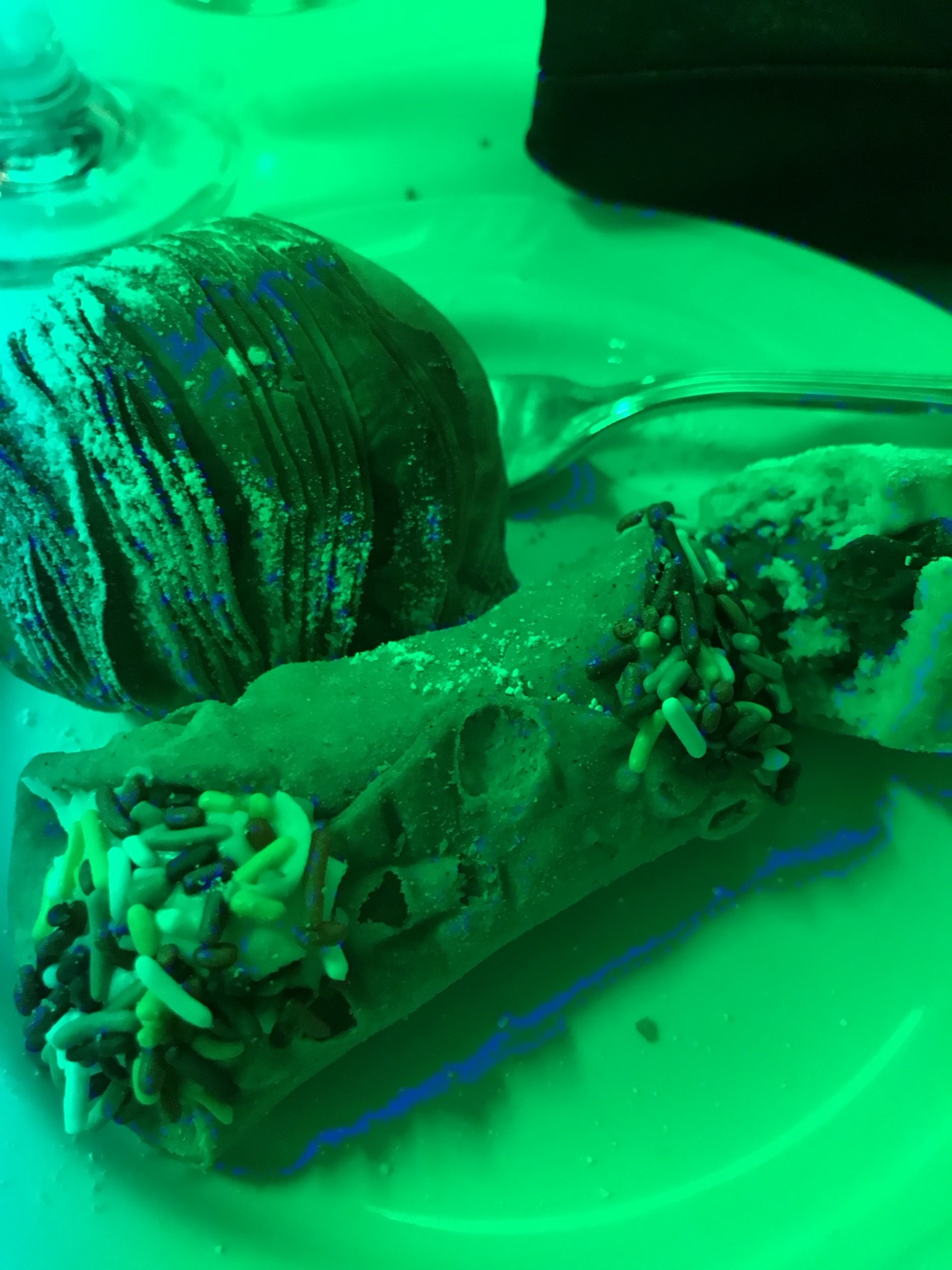 A chocolate-covered pastry and another dessert on a plate under vivid green party lighting.