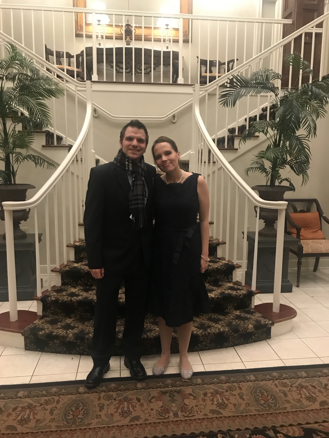Two wedding guests smiling together in formal clothes beneath an elegant curved staircase.
