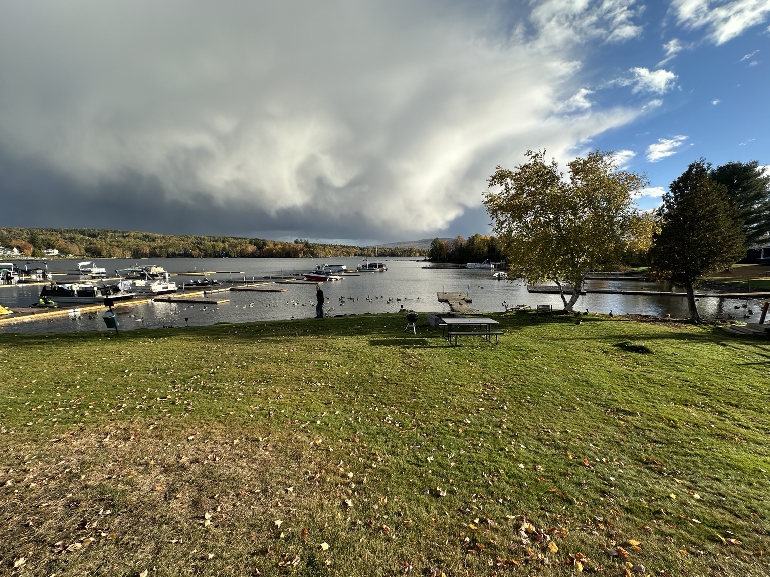 A wide Rangeley waterfront view with a grassy lawn, picnic table, docks and fall trees beneath dramatic clouds and a patch of blue sky.