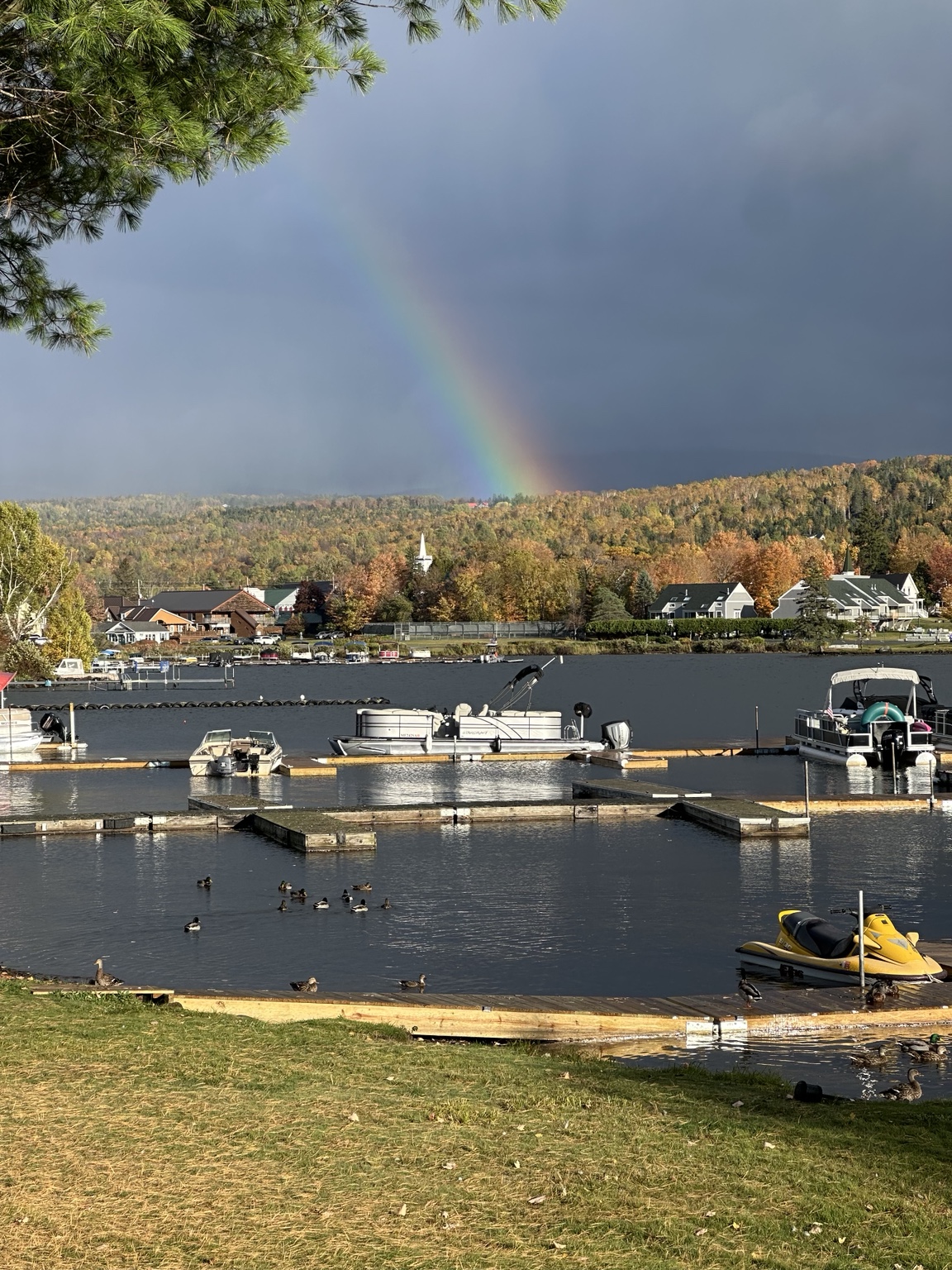A rainbow beneath dark clouds over an autumn hillside, village buildings and boats at a Rangeley waterfront.