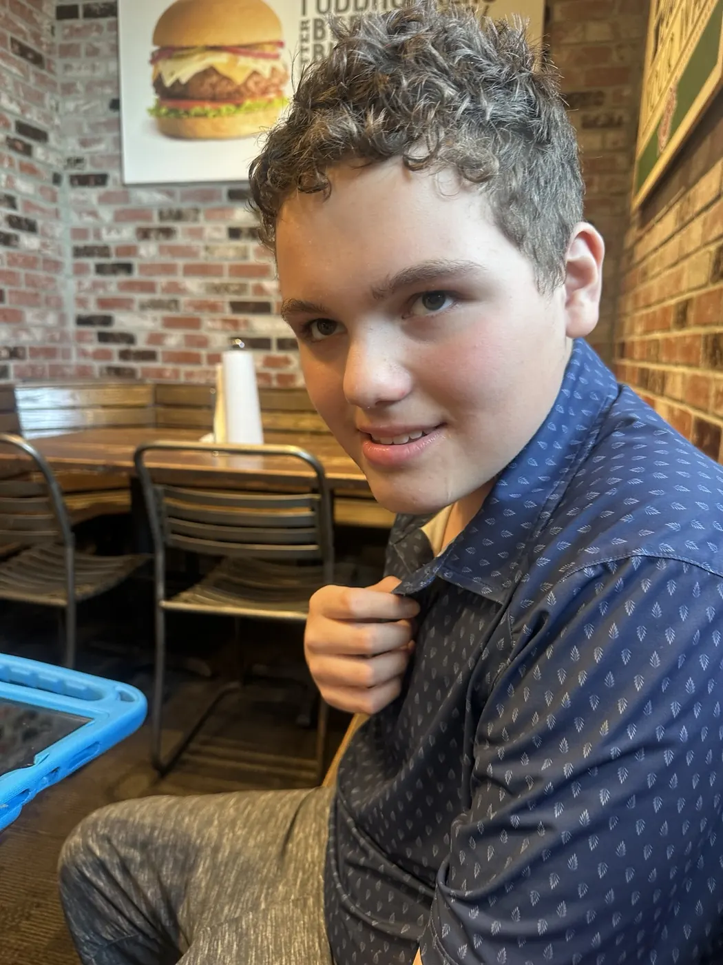 James smiling in a navy patterned shirt while seated at Fuddruckers, with brick walls and restaurant tables behind him.