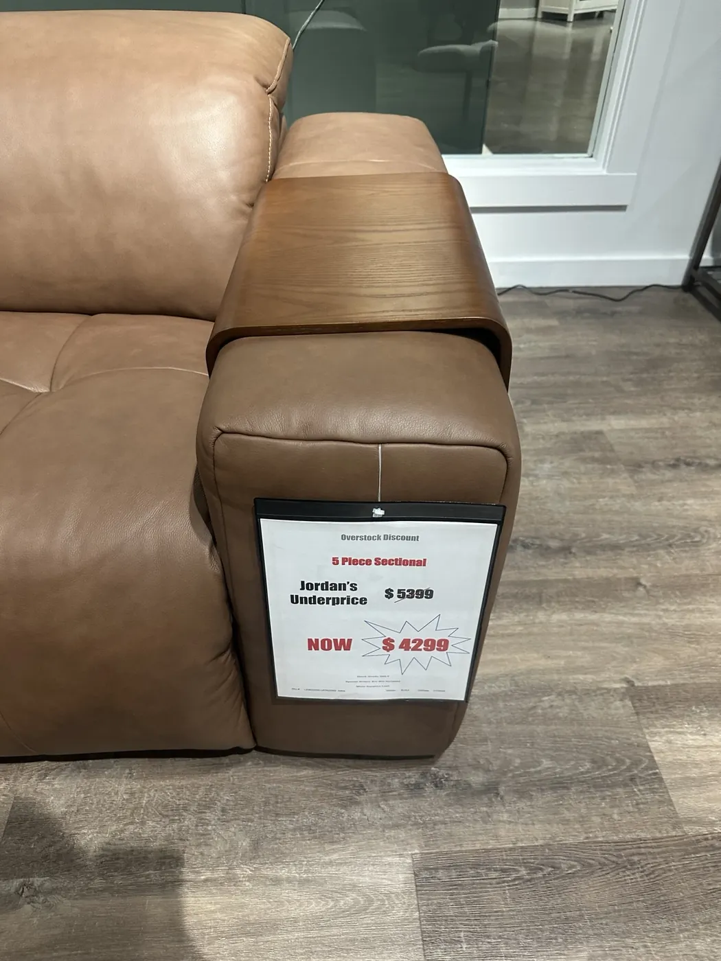 A tan leather sectional couch at Jordan’s Furniture, with a dark wooden tray-style armrest and a clearance price tag.