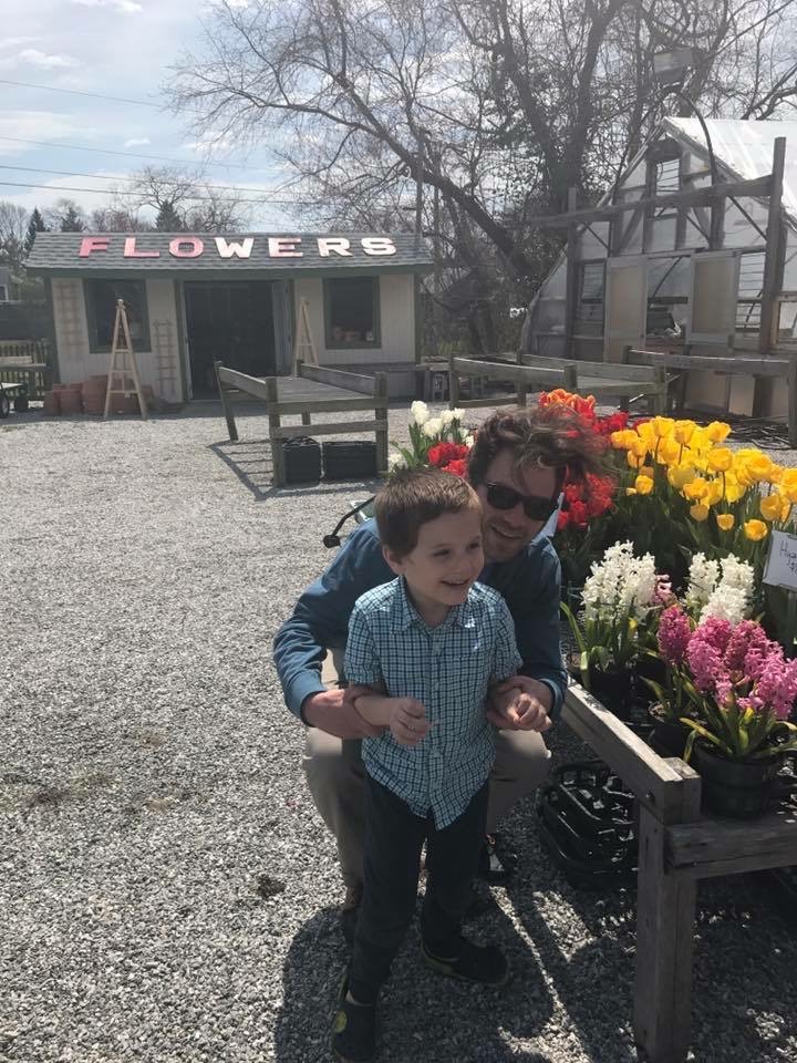 Young James smiles with Michael crouching behind him beside yellow tulips and colorful hyacinths, with a small FLOWERS shed and greenhouse behind them.