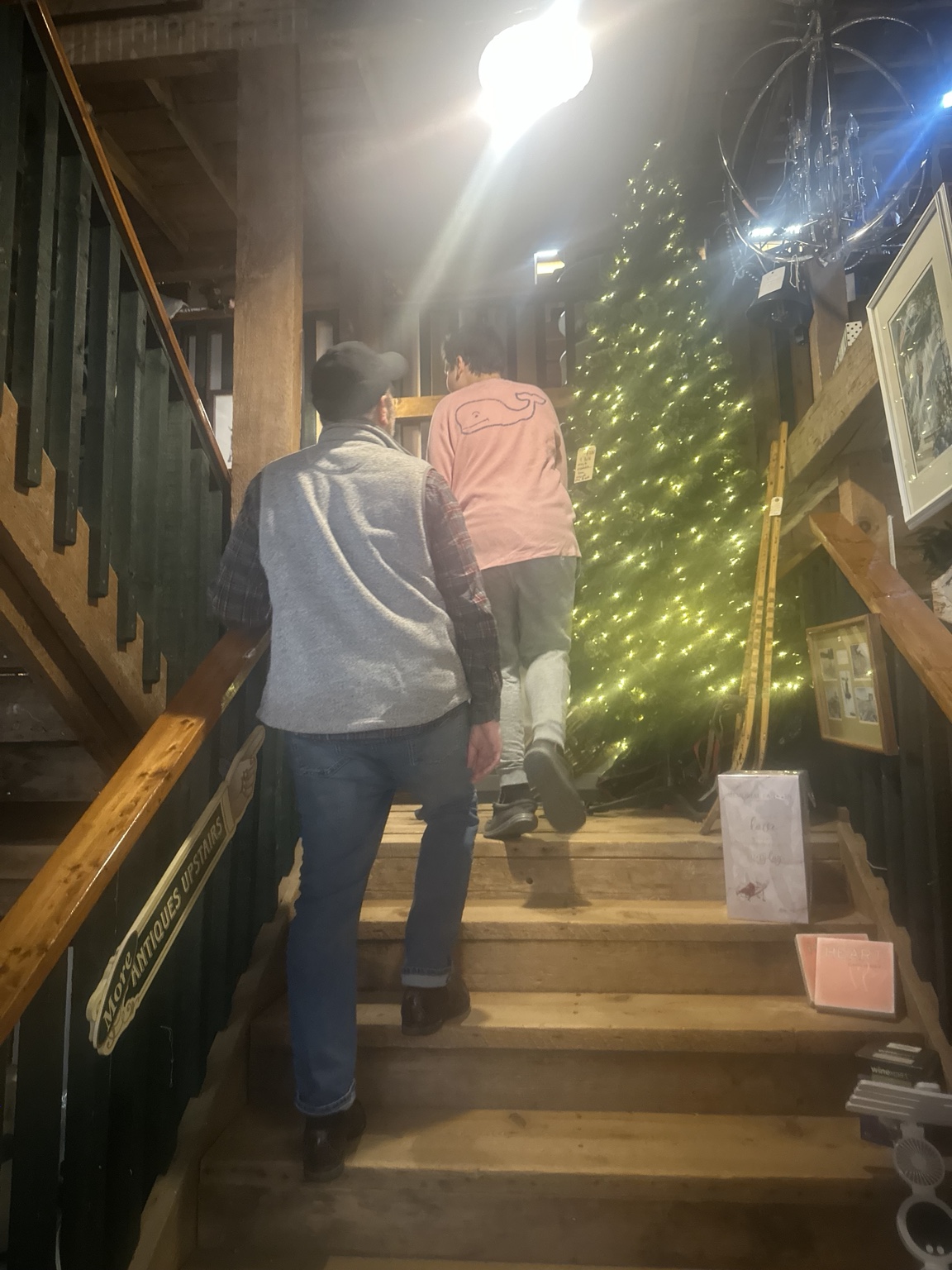 Two family members climb the wooden staircase toward upstairs antiques, beside handrails, framed art and a lighted tree.