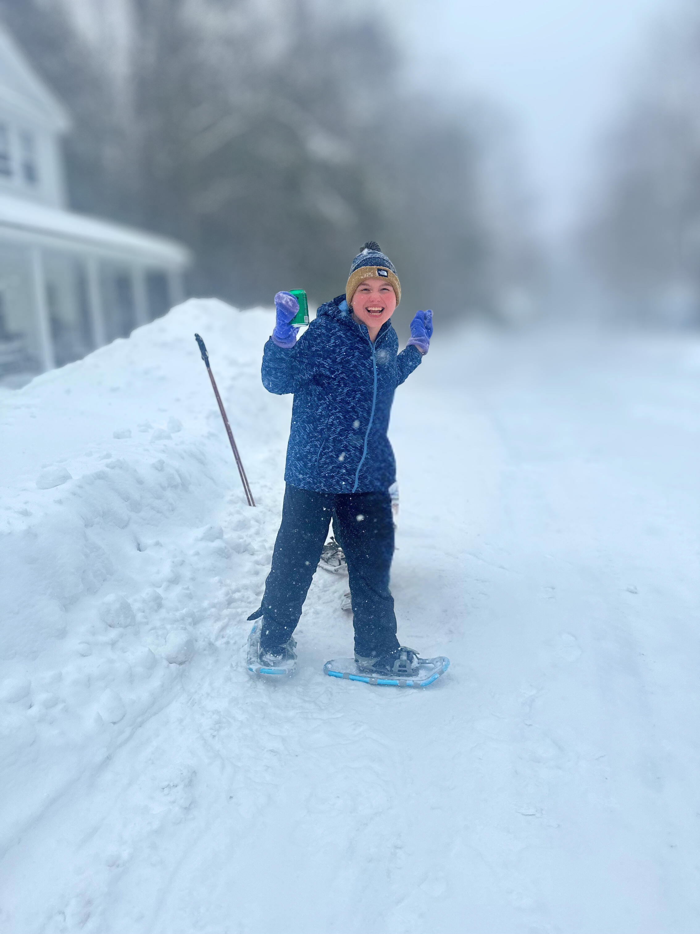 A smiling snowshoer in a navy jacket and knitted hat stands in fresh snow outside the family home, with ski poles nearby.