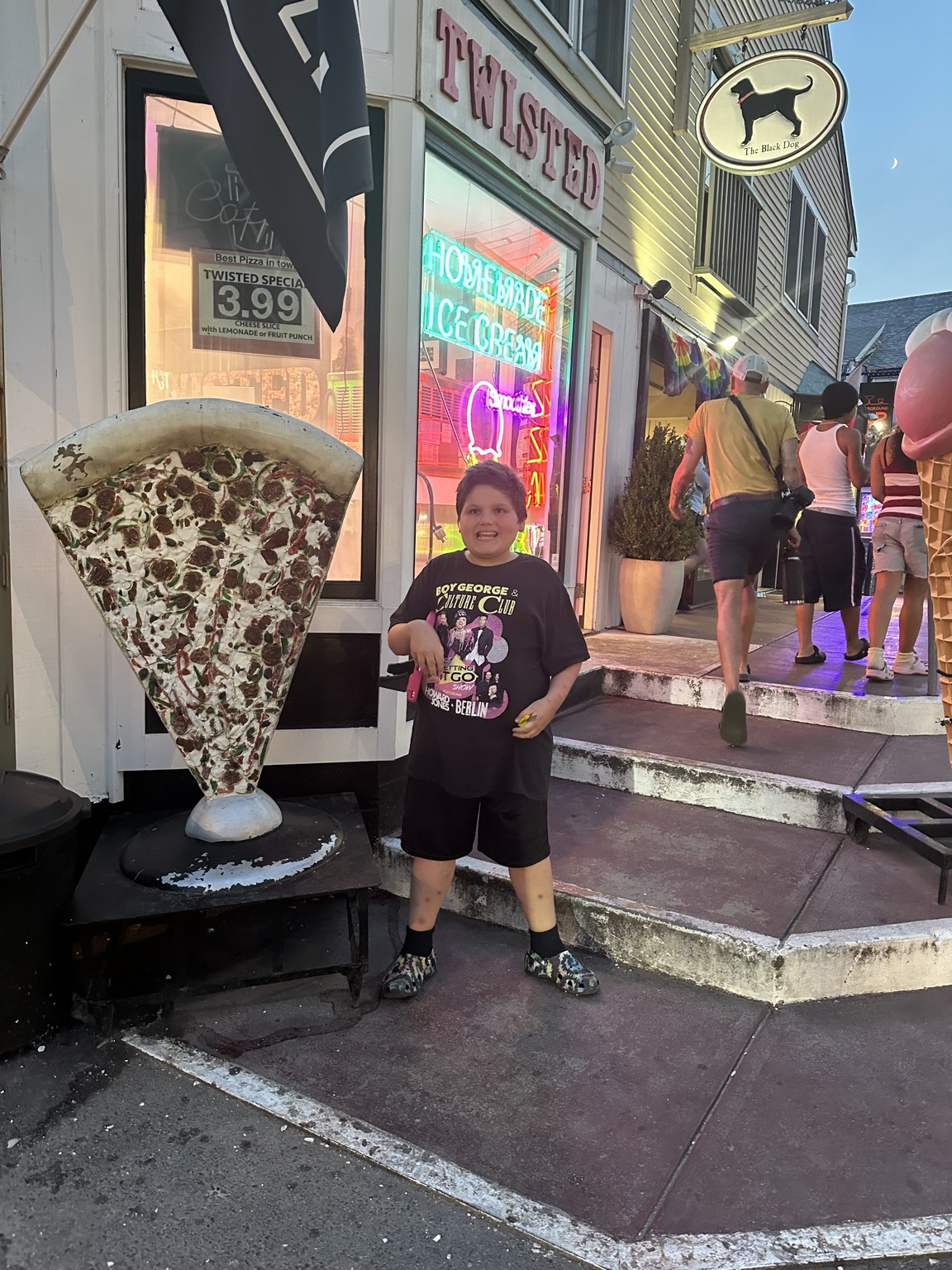 A smiling child stands beside the giant pizza-slice display outside Twisted in Provincetown, with neon signs glowing in the windows.