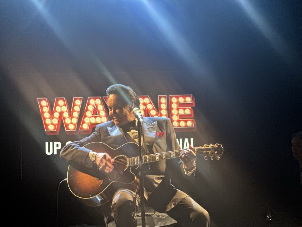 Wayne Newton seated onstage in a blue-gray suit, playing an acoustic guitar beneath the Wayne sign.
