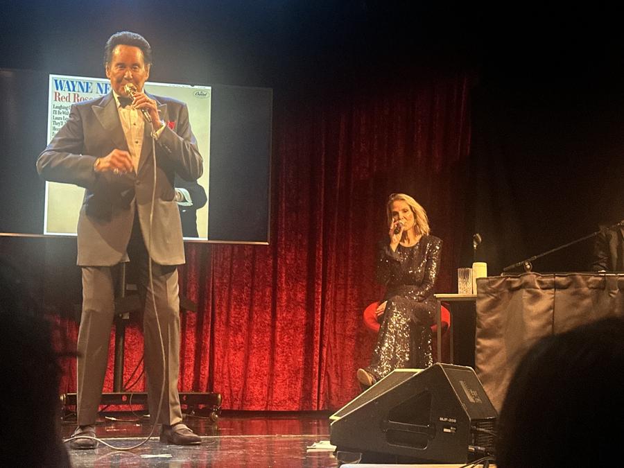 Wayne Newton singing into a microphone while a woman in a dark sequined dress sits at the side of the stage.