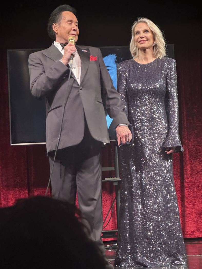 Wayne Newton onstage beside a guest performer in a sparkling gown, framed by the red curtains of Bugsy’s Cabaret.