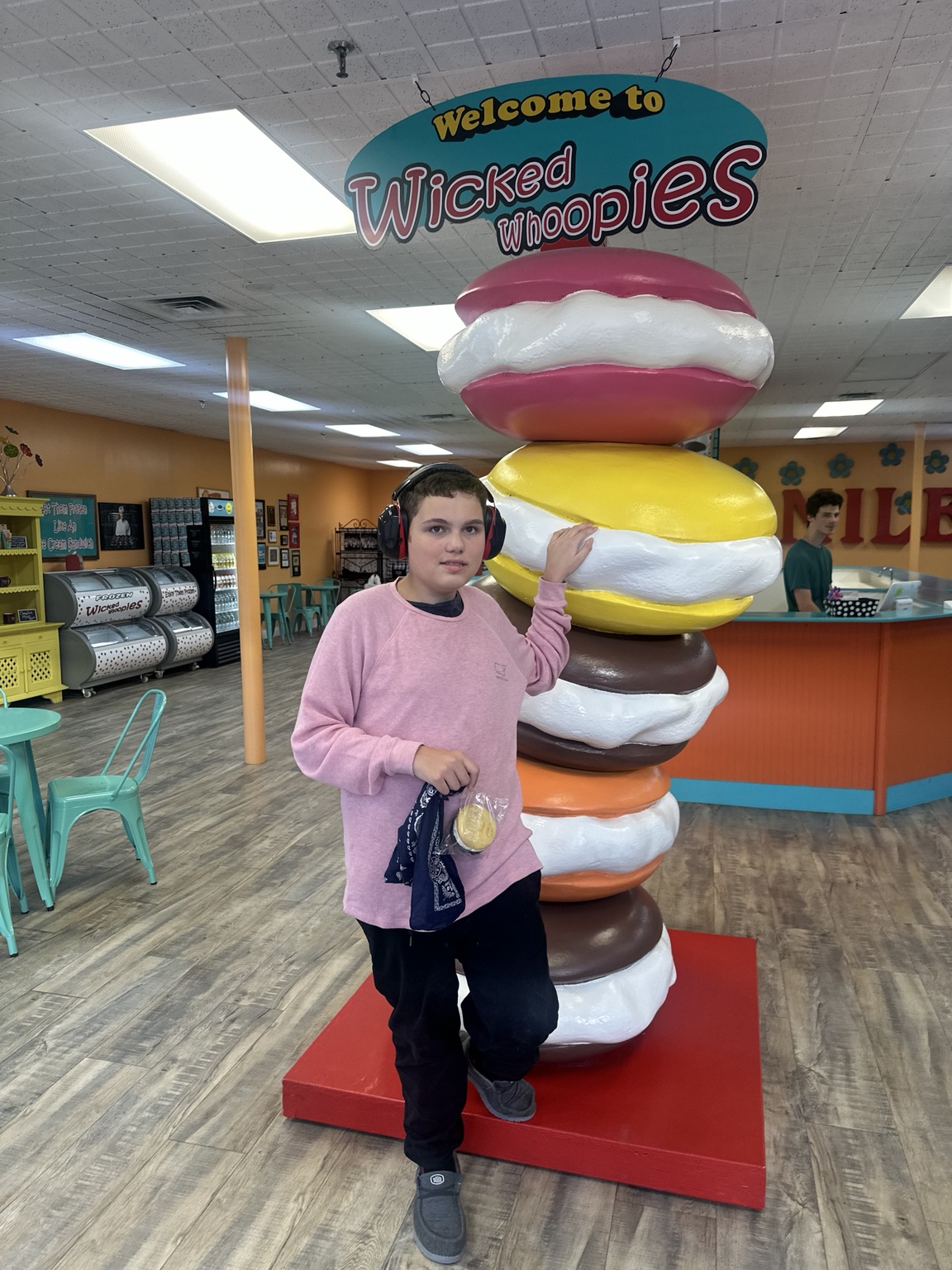 James wearing hearing protection and holding a wrapped treat beside a towering pink, yellow, chocolate and orange whoopie-pie display.